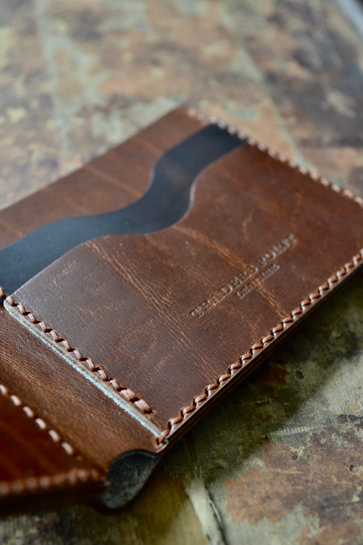 Close view along the edge of an open brown wallet, the saddle stitching running down the burnished edge and the Traders Point Leather mark pressed into the panel beside it