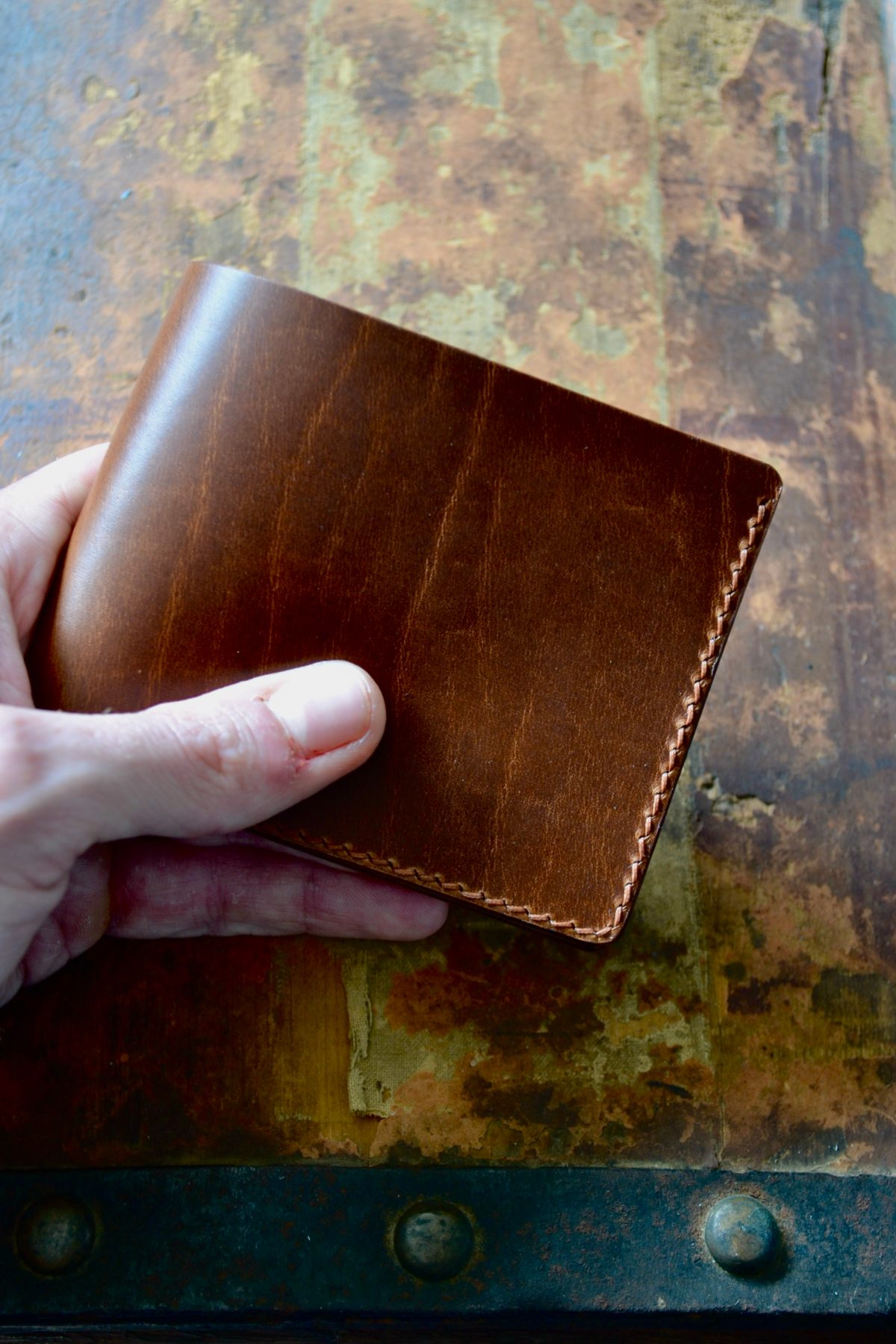 A hand holding a closed brown bifold wallet over a studded metal trunk, the stitching running down its outside edge